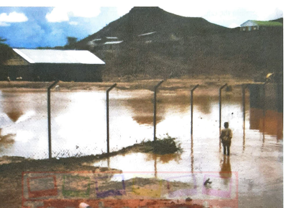 Photograph: a flooded area — muddy water covering the ground around a fence and a building, a person standing in the water, hills in the background.